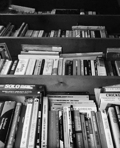 A high-contrast, black and white photograph of a densely packed bookshelf. The rows of books are layered with a mix of leaning and stacked spines, creating a textured, monolithic wall of literature. The top shelves fall into deep, dramatic shadows, evoking a sense of an endless "Internal Library."