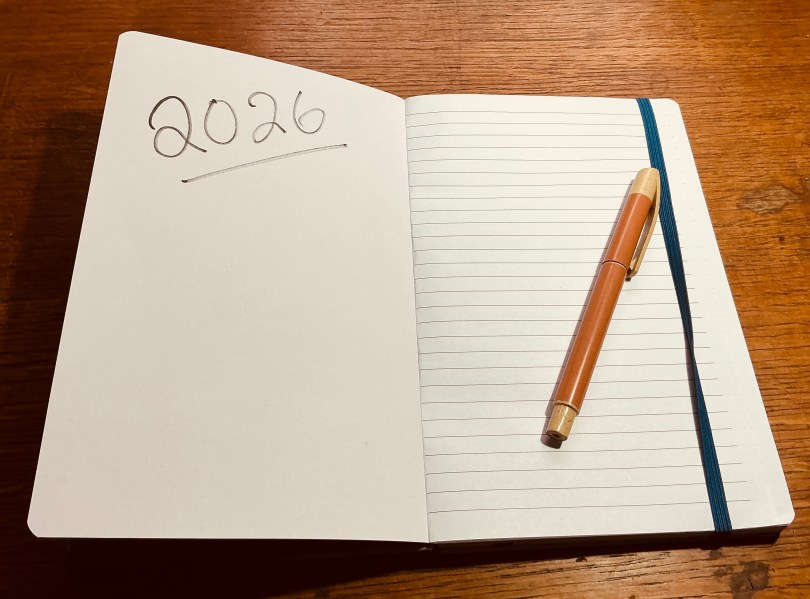 A top-down photo of an open notebook on a wooden desk. On the left blank page, '2026' is handwritten in ink. On the right lined page, a tan and orange pen rests next to a blue elastic bookmark.